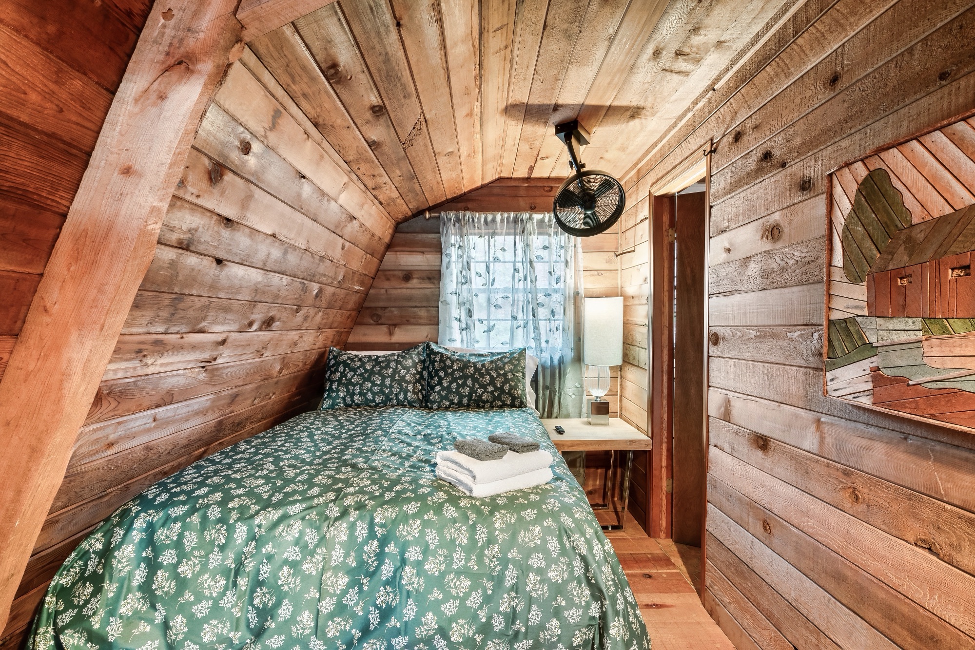 Loft bedroom at Rustic Retreat tucked under the sloped A frame roofline, with a bed in green floral bedding and a wall mounted fan